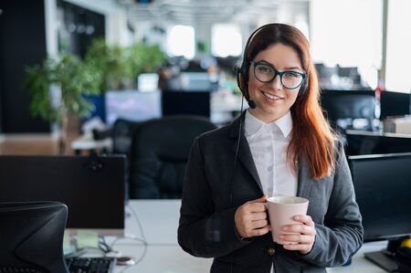 Happy office manager wearing glasses and headset holding mug. smiling female call center employee is...