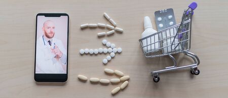 Online medicine. Remote consultation of a pharmacist doctor on a smartphone. Top view on the smartphone arrows laid out of tablets mini shopping cart and medicine lying on the table.の写真素材