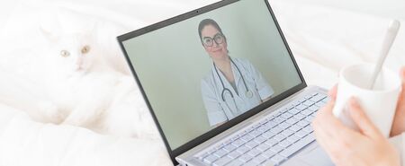 A faceless woman drinks an antipyretic and watches a video from a doctor recommendation. Online doctor consultation on a webcam without leaving your home. The cat lies in bed with a sick housewife.の写真素材