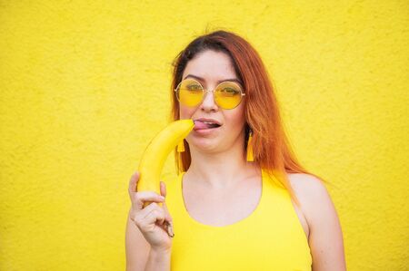A beautiful red-haired woman in a yellow dress and sunglasses shows her tongue and and licks a banana. Portrait of a fantasizing girl with ripe fruit.の写真素材
