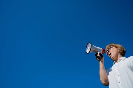 Angry senior woman in glasses shouting into a megaphone on the outside. Annoyed pensioner yells into...
