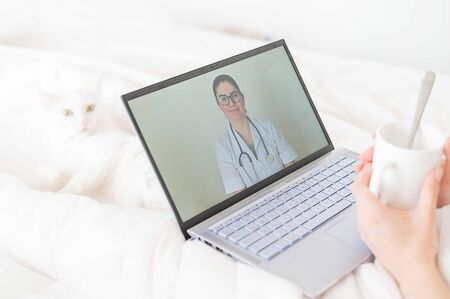 A faceless woman drinks an antipyretic and watches a video from a doctor recommendation. Online doctor consultation on a webcam without leaving your home. The cat lies in bed with a sick housewife.の写真素材