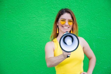 Happy woman in a yellow dress glasses and earrings on a green background shouting into a megaphone. Portrait of a girl holding a loudspeaker. Lady with a perfect snow-white smile.の写真素材