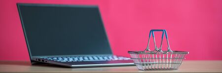 A mini shopping basket is standing in front of a laptop on a pink background. Online shopping...