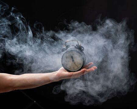 A woman holds an alarm clock in a studio full of smoke. White fog enveloped a round retro mechanical watch.の写真素材