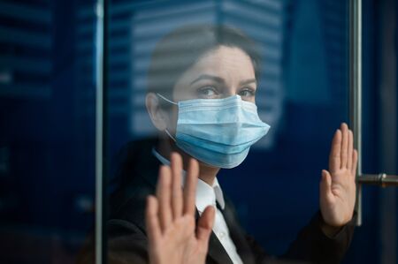 A sad quarantined woman in a business suit leaned against the window. Caucasian girl in a mask on...