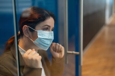 A girl in a medical mask is isolated due to biological hazard. The business woman in the suit is quarantined and leaned against the glass door with sadness. The epidemic of the virus.の写真素材