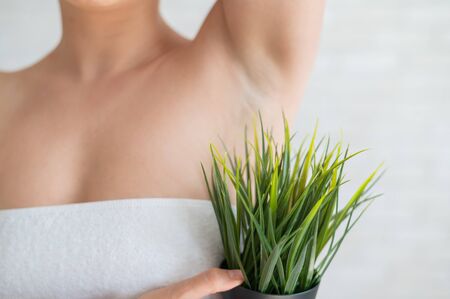 Faceless woman in a white towel holds a pot with a plant. An unrecognizable girl in a green bush imitates the vegetation of armpit hair. Depilation area. Female hair removal.の写真素材