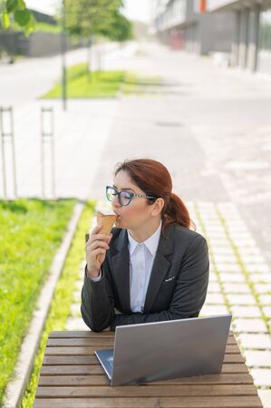 A smiling woman works remotely on a laptop and enjoys eating an ice cream cone while sitting at a wooden table. Portrait of a girl in a business suit for a break in a street cafe on a hot summer day.の写真素材