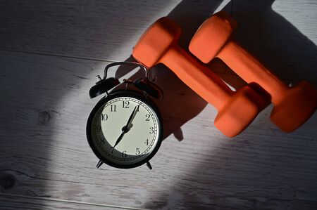 Its time to do sports early in the morning. Top view on alarm clock and orange dumbbells on a wooden floor.の写真素材