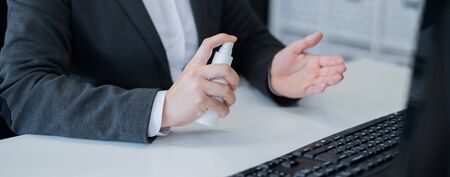 Close-up of female hands office manager at the desk using an antiseptic to disinfect. A woman in a suit treats hands with a sanitizer working at a computer. Mask on the keyboard.の写真素材