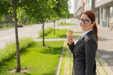 Business woman walks outdoors and enjoys vanilla ice-cream. Happy girl in a suit eats an ice cream cone on a hot sunny summer day. Lunch break of an office employee.の写真素材