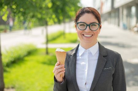 businesswoman is walking outdoors and eating vanilla icecream on a sunny summer day. Portrait of a smiling girl in a white shirt and a jacket with an ice cream cone. Lunch break of an office employee.の写真素材