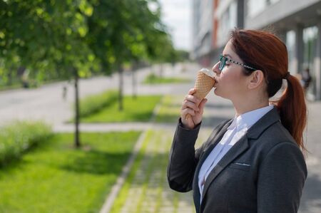 A business woman walks outdoors and enjoys eating vanilla ice-cream on a sunny summer day. Portrait of a smiling girl in a white shirt and a jacket with an ice cream cone.の写真素材