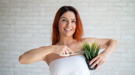 A smiling red-haired woman in a white terry towel cuts a green plant in a pot with scissors. Imitation of armpit depilation. Hair removal concept. Procedure for hair removal of unwanted vegetation.の写真素材