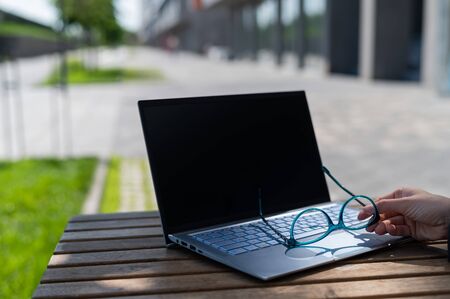 Close-up of a female hand holds glasses near a laptop on a wooden table in a summer cafe. Freelancer workplace. A woman remotely working outdoors.の写真素材