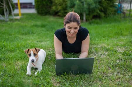 A red-haired woman lies on the green grass in a park with her own dog. The girl maintains a social distance and types on a laptop in the fresh air. A student is studying remotely on a computer.の写真素材