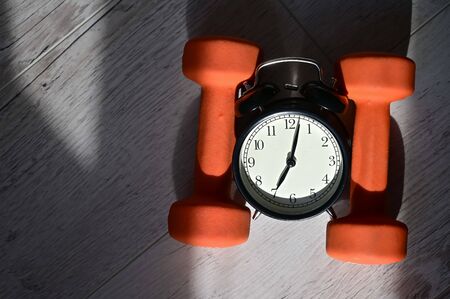 Its time to do sports early in the morning. Top view on alarm clock and orange dumbbells on a wooden...