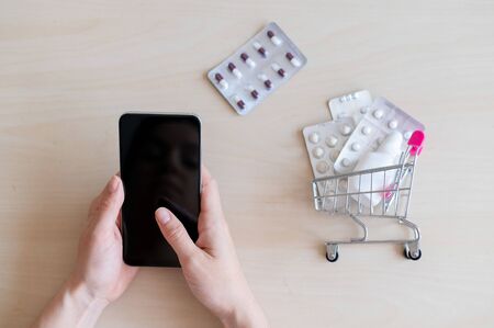 The concept of remote selling drugs on a mobile application. online pharmacy. A smartphone with a black screen in female hands and a mini trolley with tablet blisters.の写真素材