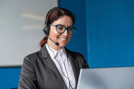 Friendly woman wearing a headset is having an online conversation on a laptop in a conference room. Female manager communicates with clients via video call. Remote business negotiations in quarantine.の写真素材