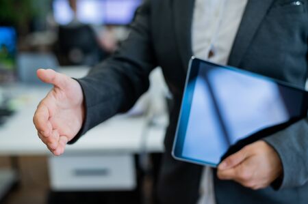 Business woman in a jacket and white shirt holding out a hand for a greeting. A faceless business woman holds a digital tablet and shakes her hand as an agreement on a successful transaction.の写真素材