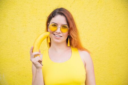 Portrait of a beautiful red-haired woman in a yellow dress and sunglasses holds a banana near...