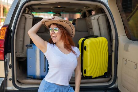 Happy red-haired woman in a cowboy hat and sunglasses going on a car trip around the country. Girl...
