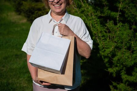 Happy elderly woman in glasses holds an eco friendly package while walking in the park. A delighted old lady stands with purchases in paper bags outdoors.の写真素材