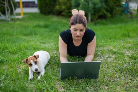 A red-haired woman lies on the green grass in a park with her own dog. The girl maintains a social distance and types on a laptop in the fresh air. A student is studying remotely on a computer.の写真素材
