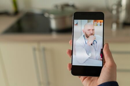 A serious bearded doctor communicates with a patient via a video call on a smartphone. Online...