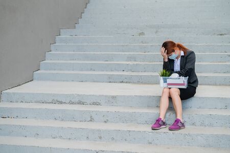 A woman with a box of personal belongings sits on the stairs outside and folded her hands in despair on head. Female employee in medical mask fired due to financial crisis in coronovirus. Unemploymentの写真素材