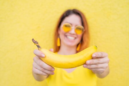 Beautiful red-haired woman in sunglasses holds a banana and fools around. Attractive European girl in a dress posing on a yellow background charmingly smiles.の写真素材