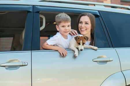 Beautiful caucasian woman travels with a child and a dog. Mom and son leaned out of the car window in an embrace with a puppy of Jack Russell Terrier. Happy family go on a car trip.の写真素材