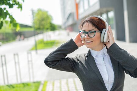 Portrait of a smiling woman in a business suit listening to music in headphones outdoors. Female office employee holds hands on earphones enjoying song and dancing on the street.の写真素材