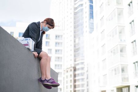 A depressed woman in a medical mask sits on the edge of the roof with a box of personal items from the desktop. An unemployed girl alone wants to commit suicide. Bankruptcy of the company.の写真素材