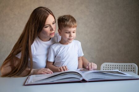 A middle-aged Caucasian woman hugs a little son and shows a photo album. A little boy sits on his...