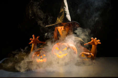 An ominous witch in a hat conjures over a jack-o-lantern. Traditional halloween characters. Mystical fog creeps over pumpkins with carved terrible faces. An evil witch casts a spell.の写真素材