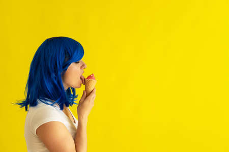 A beautiful woman with blue hair stands in profile and licks a horn of raspberry ice cream on a yellow background. The girl in the wig enjoys eating gelato in a waffle cup. Summer cooling dessert.の写真素材