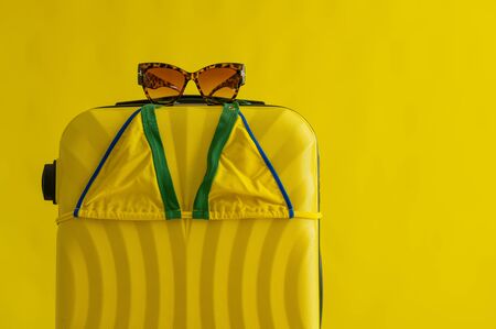 Yellow suitcase on wheels with tiger sunglasses and a mini bikini on a yellow background in the studio. Set for a beach vacation. Copy space. No people. Summer vacation at sea.の写真素材