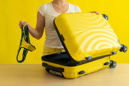 Faceless woman packs a mini bikini in a suitcase on a yellow background. The girl is going on a trip to the sea and puts a swimsuit in a bag. Ready for vacation. Cropped. Air flight journey conceptの写真素材