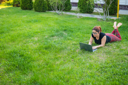 Woman is shopping on a laptop while lying on her stomach on green grass in a park. A student with two glasses is studying at personal wireless computers while lying on the lawn. Online shopping.の写真素材