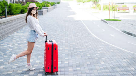 A beautiful young Caucasian woman in a medical mask travels with a red suitcase through an empty city. The girl goes on vacation with a large luggage bag during the coronavirus epidemic.の写真素材