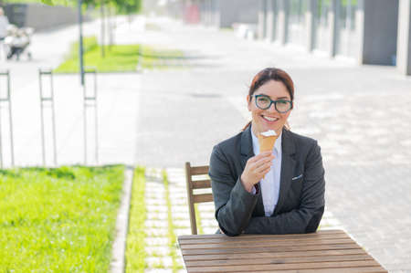 Smiling woman enjoys eating an ice cream cone while sitting at a wooden table outdoors. Portrait of a girl in a business suit for a break in a street cafe on a hot summer day.の写真素材