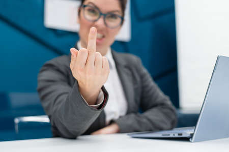 Angry business woman in a suit shows a middle finger while sitting at a desk. Annoyed female office worker showing a fuck you gesture. Professional burnout.の写真素材