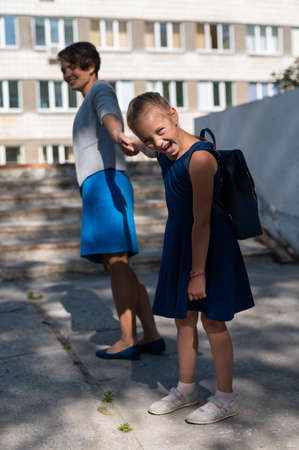 A little unhappy girl with a backpack pulls her mother by the hand does not want to go to school. A woman leads a resisting daughter to school. The conflict of generations.の写真素材