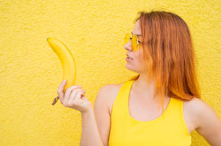 Portrait of caucasian redhead woman in sunglasses holding a banana in her hands. Beautiful seductive girl posing on a yellow background.の写真素材