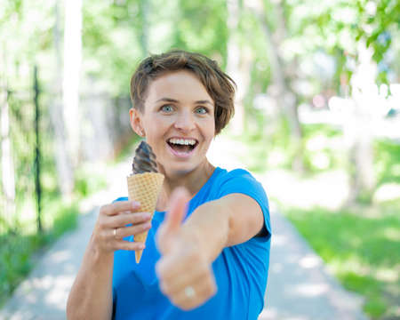 Charming Caucasian woman with a short haircut smiles and eats an ice cream cone and shows a thumb....