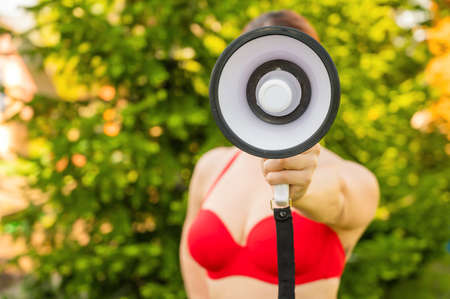 Close-up portrait of a woman in a red bikini holding a megaphone. A girl in a swimsuit makes an announcement on the loudspeaker on the background of a green treeの写真素材