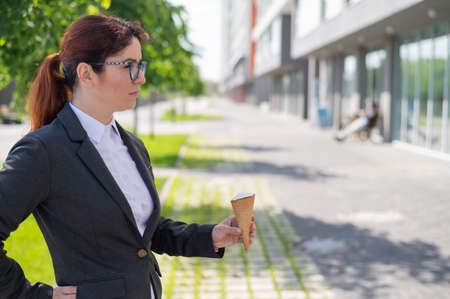 A business woman walks outdoors and enjoys eating vanilla ice-cream on a sunny summer day. Portrait of a smiling girl in a white shirt and a jacket with an ice cream cone.の写真素材
