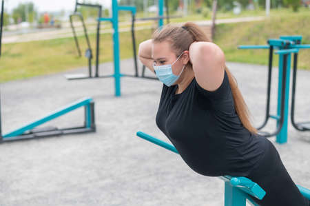 Overweight young woman in a protective mask plays sports on a playground. The girl does exercises...